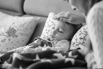 Child laying under a blanket, mom's hand placed on child's forehead