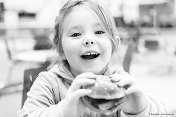 young girl around the age of 5 with a burger in her hands as she smiles at the camera