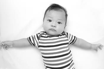 A young infant is laying down with arms sprawled out. The infant is wearing a striped onesie.