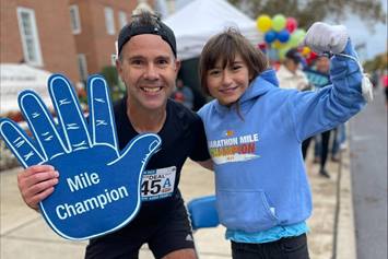 A runner holds a "Mile Champion" sign in one hand with his other arm wrapped around a smiling kid.