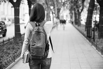A teenage girl has her back to the camera. She's wearing a back pack and is walking on a path in a tree-lined public area.